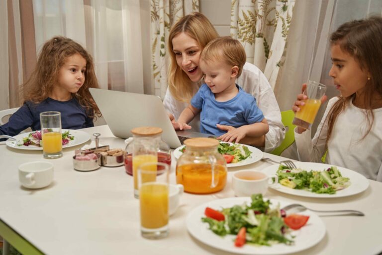 a woman sits at a dinner table with three children looking at a laptop